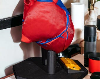 May include: A 3D printed anatomical heart model featuring a red main body with blue vascular details and a red top section. The model is mounted on a black octagonal base with a central support pillar and a small yellow rectangular nameplate.
