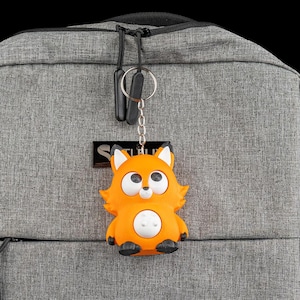Könnte beinhalten: Ein orangefarbener Fuchsschlüsselanhänger mit weißen Akzenten und schwarzen Füßen hängt an einer silbernen Kette. Der Fuchs hat große, cartoonartige Augen und einen weißen Bauch. Der Schlüsselanhänger ist an einem grauen Rucksack befestigt.