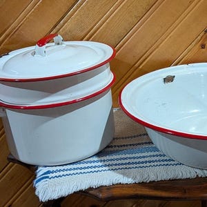 May include: A collection of vintage white enamelware kitchen items with red trim. Includes a double boiler with red handles, a large bowl, and a rectangular dish. The items are arranged on a wooden table with a blue and white striped cloth.