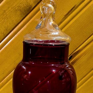 Könnte beinhalten: Ein rubinrotes Apothekerglas mit einem Deckel aus klarem Glas. Das Glas hat einen bauchigen Körper und steht auf einem Stiel und einer Basis aus klarem Glas. Das Glas ist mit einer dunkelroten Flüssigkeit gefüllt.