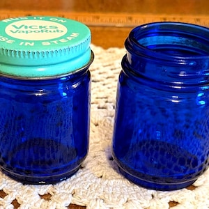 Könnte beinhalten: Zwei kobaltblaue Glasbehälter. Einer hat einen türkisfarbenen Deckel mit dem Text "RUN IT ON VICKS VapoRub USE IN STEAM". Die Gläser stehen auf einer weißen Häkeloberfläche.