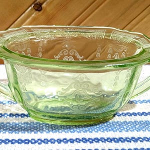 May include: A light green, vintage glass bowl with two handles. The bowl has a decorative etched pattern around the rim and on the sides. The bowl is sitting on a blue and white striped cloth.