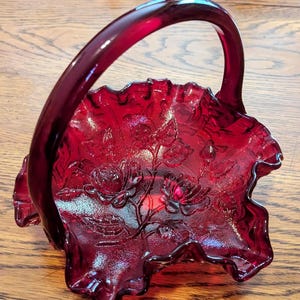 May include: Ruby red glass basket with a handle and a ruffled edge. The basket features an embossed floral design with roses and leaves. The handle is arched and smooth. The basket is sitting on a wooden surface.