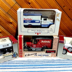 May include: Three die-cast metal toy truck banks in original packaging. The top box features a 1931 True Value delivery truck. The bottom left box contains a black and white Master Mechanic truck, and the bottom right box holds a red and white Tru-Test truck.