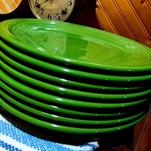 May include: A stack of eight bright green oval-shaped ceramic plates. The plates are stacked on top of each other, showcasing their glossy finish. The plates are part of a larger set of colorful dishware.