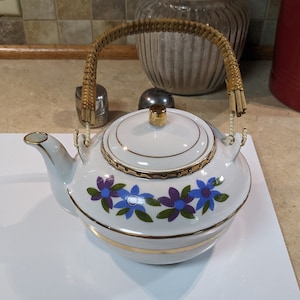 May include: White teapot with a gold trim and blue floral design. The teapot has a woven handle.