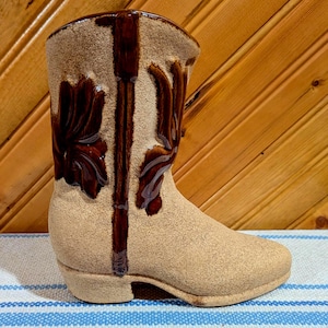 May include: A brown ceramic cowboy boot vase with a textured surface and brown floral designs. The boot is sitting on a blue and white striped surface.
