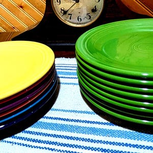 May include: A stack of bright green ceramic plates next to a stack of yellow, purple, red, and blue plates. The plates are on a blue and white striped cloth. A clock and wood paneling are in the background.