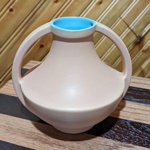 May include: A light pink ceramic vase with two handles. The vase has a light blue rim and a rounded body.
