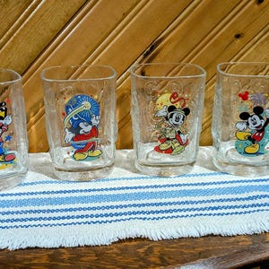 Puede incluir: Cuatro vasos de vidrio transparente con personajes de Mickey Mouse. Cada vaso presenta un personaje de Mickey Mouse diferente con un diseño único. Los vasos están sobre una tela a rayas azules y blancas.