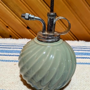 May include: A sage green glass plant mister with a spherical, swirled body and a metallic pump mechanism. The device features a chrome-finished nozzle, a circular handle, and a white knob at the top of the vertical plunger for manual operation.