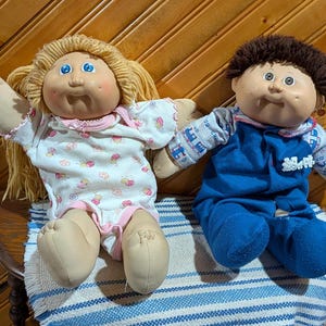 May include: Two vintage Cabbage Patch Kids dolls. One doll has blonde yarn hair and wears a white onesie with pink trim and a floral pattern. The other doll has brown hair and wears a blue jumpsuit with a patterned shirt.