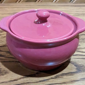 以下が含まれることがあります： 蓋付きのピンクのセラミック製のキャセロール。丸みを帯びた形と滑らかで光沢のある仕上げです。