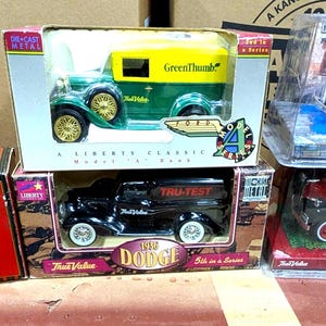 May include: A collection of vintage toy trucks in their original boxes. The trucks are various colors, including red, green, and black, with detailed designs and branding. Some boxes feature clear plastic windows.