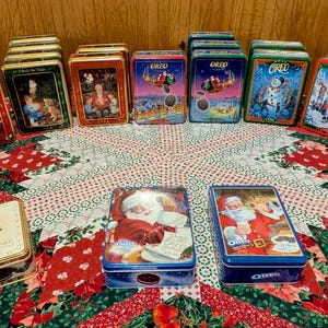 May include: A collection of vintage Christmas-themed tins, featuring Santa Claus, winter scenes, and festive designs. The tins are arranged on a patterned tablecloth, showcasing a variety of shapes and sizes, with a mix of red, gold, and blue colors.