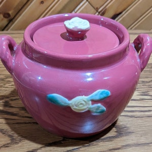 May include: A pink ceramic jar with a lid and two handles. The jar has a floral design with a white rose and green leaves.