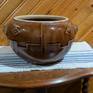 May include: A brown ceramic pot with a decorative design and two handles. The pot sits on a wooden table covered with a white and blue striped cloth. The background is a wooden wall.