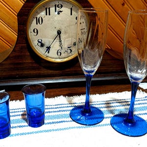 May include: Two clear glass champagne flutes with blue stems and bases, accompanied by two small, solid blue glass shot glasses. The flutes feature a subtle vertical swirl pattern in the glass, while the shot glasses have a smooth, deep blue finish.