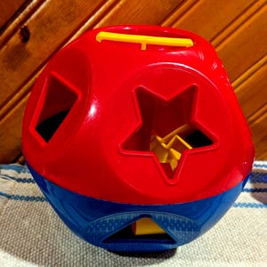 Könnte beinhalten: Ein kugelförmiges Formensortierspielzeug aus Kunststoff mit einer leuchtend roten oberen Hälfte und einer dunkelblauen unteren Hälfte. Das Spielzeug enthält verschiedene ausgestanzte Formen wie einen Stern und ein Quadrat sowie einen gelben Griff.