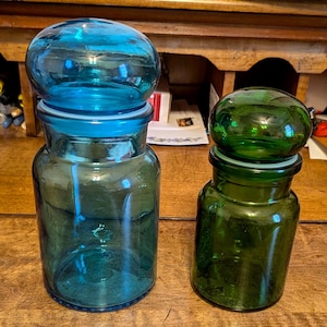 May include: Two glass apothecary jars with round lids. One jar is teal blue, and the other is green. Both jars have a cylindrical shape with a wide base and a matching lid. The jars are sitting on a wooden surface.