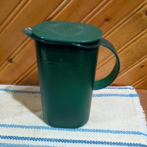 Può includere: Una brocca verde scuro con coperchio e manico abbinati. La brocca è realizzata in un materiale solido e poggia su un panno a righe bianche e blu. Lo sfondo è una parete di legno.
