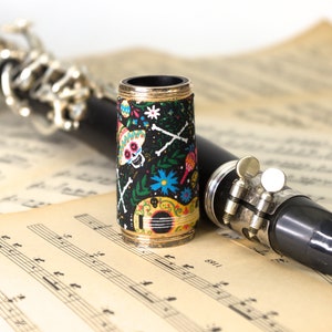 Puede incluir: Un clarinete negro con un cañón decorativo con estampado de calaveras y flores, que descansa sobre una partitura musical.