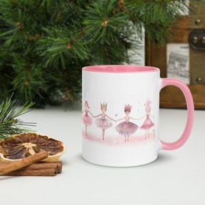 May include: A white ceramic mug with a pink rim and handle. The mug features a watercolor illustration of five ballerinas in pink tutus holding hands. The ballerinas are wearing crowns and have pink hair.