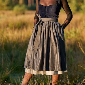 Op de afbeelding: Een vrouw in een donkerblauwe en grijze dirndl-jurk met een zwart lint. Ze staat in een veld met hoog gras.