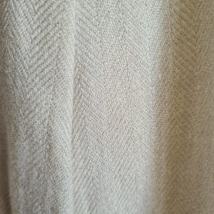 Pode incluir: Close-up de um tecido bege com um padrão de tecido em espinha de peixe. A textura parece macia e ligeiramente irregular, com variações sutis de cor. A trama cria um padrão diagonal na superfície.