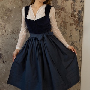 Op de afbeelding: Donkerblauwe dirndljurk met een witte blouse en een donkerblauwe schort. De jurk heeft een vierkante halslijn en een strik in de taille. De tekst "DARK BLUE" staat onderaan de afbeelding.