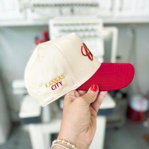 May include: A white baseball cap with a red brim. The cap has "KANSAS CITY" embroidered in gold on the front and a red "C" embroidered on the side.