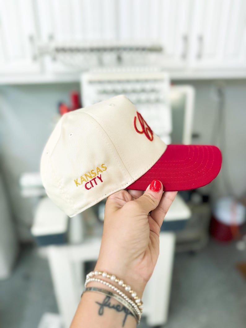 May include: A white baseball cap with a red brim. The cap has "KANSAS CITY" embroidered in gold on the front and a red "C" embroidered on the side.
