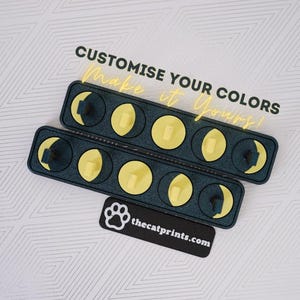 Puede incluir: Dos organizadores de color verde azulado oscuro con inserciones circulares amarillas y pequeños objetos rectangulares de color claro. El texto "CUSTOMISE YOUR COLORS Make it Yours!" se muestra en amarillo. También es visible una etiqueta negra con una huella de pata y thecatprints.com.
