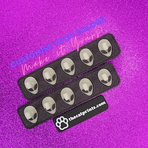 May include: Alien-themed press-on nails arranged in rows on a black tray, set against a sparkly purple background. The nails feature a white base with a gray alien face design. The text "CUSTOMISE YOUR COLORS Make it Yours!" is visible.