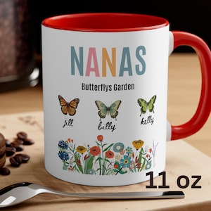 May include: White ceramic mug with a red rim and handle. The mug has a floral design with butterflies and the text "NANAS Butterflys Garden" with the names "Jill, Billy, Kelly" written below the butterflies. The mug is 11 oz.