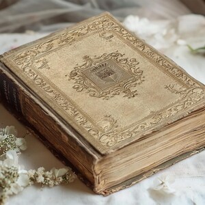 May include: A vintage, leather-bound book with a gold-embossed design on the cover. The book is closed and lying on a white fabric surface.