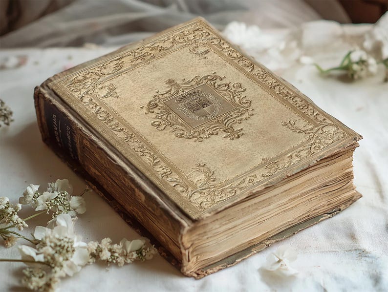 May include: A vintage, leather-bound book with a gold-embossed design on the cover. The book is closed and lying on a white fabric surface.