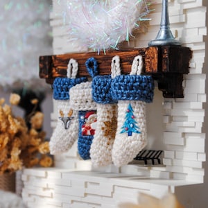 May include: Four white crocheted stockings with blue crocheted tops hang from a wooden mantel. The stockings are decorated with embroidered designs, including a reindeer, Santa Claus, a star, and a Christmas tree.