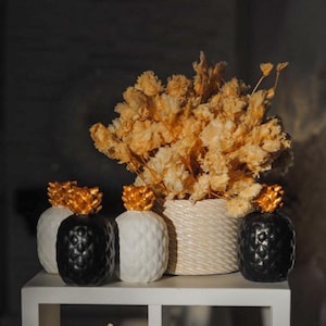 May include: White shelf unit with three drawers, decorated with black and white pineapple figurines, a white pumpkin with black polka dots, and dried flowers in a woven basket.