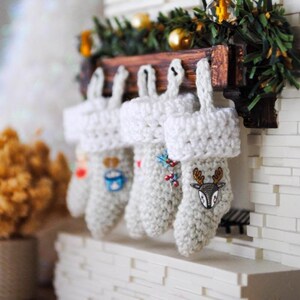 May include: Five white crocheted Christmas stockings with different embroidered designs hang on a wooden mantel above a white brick fireplace.