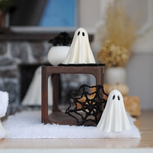 May include: Two white ghost figurines with black eyes on a wooden shelf with a black spiderweb decoration. The shelf is in front of a stone fireplace.