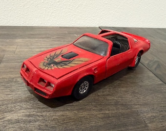 Puede incluir: Un coche de juguete rojo con un emblema de águila negro y dorado en el capó. El coche tiene ruedas negras y un alerón. Es un modelo vintage, probablemente una réplica de un coche deportivo clásico.