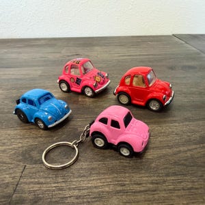 May include: Four toy cars shaped like classic beetles. One is bright blue, one is red, one is hot pink with black graphic designs and the text Hi, and one is a light pink car attached to a silver metal keyring with a circular ring attachment.