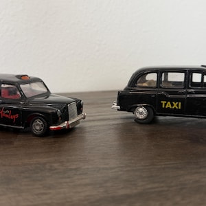 May include: Two black die-cast toy taxi cabs. The left vehicle features red interior seating and the red text Hamleys on the side. The right vehicle has the word TAXI printed in yellow on the side door. Both models have silver wheels and bumpers.