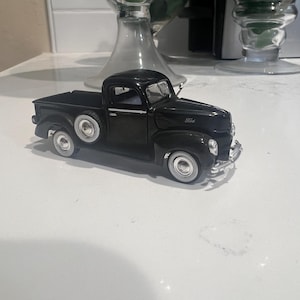 May include: A black vintage Ford pickup truck model with white-walled tires. The truck has a detailed design, including a front grill, headlights, and the Ford logo. The model is a classic collectible item.