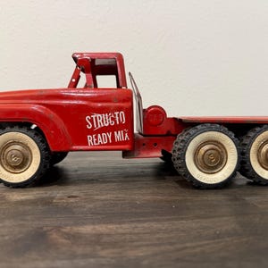 May include: A vintage red metal toy truck with the white text Structo Ready Mix printed on the side door. The truck features a cab, a flatbed rear section, and six wheels with white sidewalls and black treads. The wheels have a small S logo.
