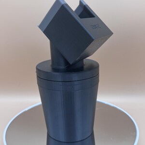 Puede incluir: Un tap&oacute;n de botella negro impreso en 3D con un dise&ntilde;o geom&eacute;trico &uacute;nico. El tap&oacute;n presenta una base cil&iacute;ndrica y una parte superior angular inclinada con una abertura rectangular. El texto "OM32" es visible en la parte superior.