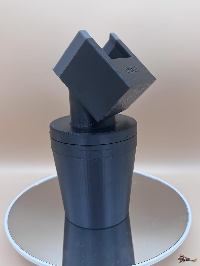 Puede incluir: Un tap&oacute;n de botella negro impreso en 3D con un dise&ntilde;o geom&eacute;trico &uacute;nico. El tap&oacute;n presenta una base cil&iacute;ndrica y una parte superior angular inclinada con una abertura rectangular. El texto "OM32" es visible en la parte superior.