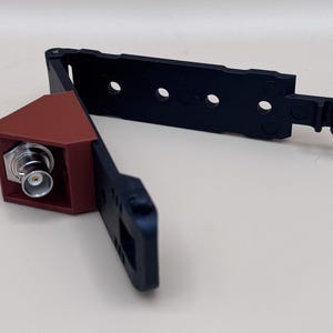 May include: A black plastic mounting bracket with a hinged design and a brown triangular housing containing a silver BNC connector. The bracket features multiple circular cutouts along its length and a hook mechanism at the end for secure attachment.
