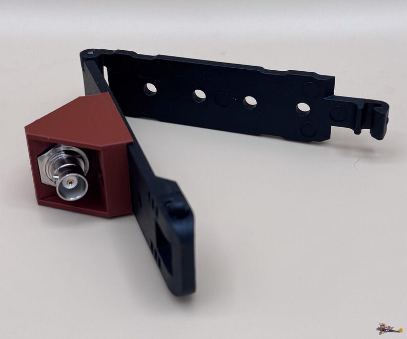 May include: A black plastic mounting bracket with a hinged design and a brown triangular housing containing a silver BNC connector. The bracket features multiple circular cutouts along its length and a hook mechanism at the end for secure attachment.